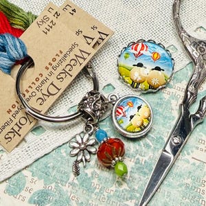 May include: A collection of cross-stitch supplies, including embroidery floss, a charm bracelet with sheep and hot air balloon pendants, and a pair of silver scissors. The floss is in shades of red, blue, and green. The charm bracelet has a floral charm and a red bead.