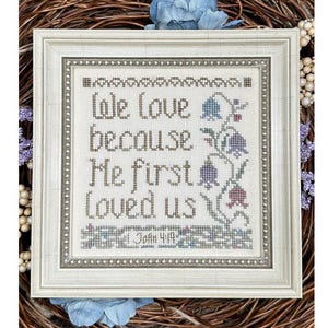 Puede incluir: Una bordadura de punto de cruz enmarcada con fondo blanco y texto marrón que dice "We love because He first loved us 1 John 4:19". La bordadura presenta un diseño floral con flores moradas, rosas y azules.
