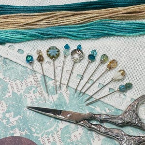 May include: Decorative pins with bead and shell designs are displayed on a patterned surface. Teal and beige threads are in the background. A pair of ornate scissors is also present. The pins are ideal for sewing.