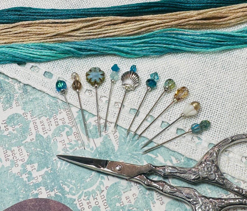 May include: Decorative pins with bead and shell designs are displayed on a patterned surface. Teal and beige threads are in the background. A pair of ornate scissors is also present. The pins are ideal for sewing.