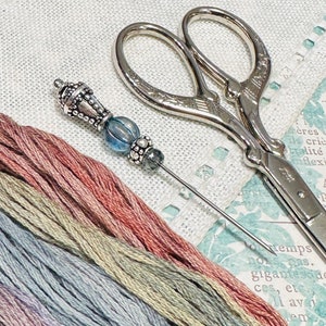May include: A silver needle threader with a blue glass bead and silver accents. The needle threader is lying on a white linen fabric with several skeins of embroidery floss in shades of blue, green, and pink. A pair of silver scissors with an ornate design is also visible in the image.