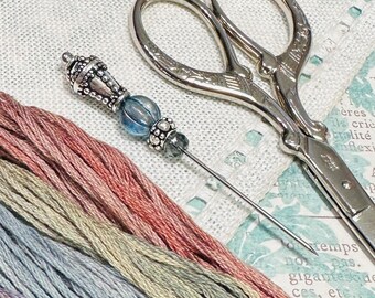 May include: A silver needle threader with a blue glass bead and silver accents. The needle threader is lying on a white linen fabric with several skeins of embroidery floss in shades of blue, green, and pink. A pair of silver scissors with an ornate design is also visible in the image.