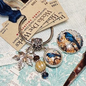 May include: A silver scissor charm with a blue bird and leaf accents. The charm is attached to a silver ring with a decorative bead. The charm is on a white fabric background with a tag that reads "Weeks Dye Works" and "Specialising in Hand Dyed Fibres".