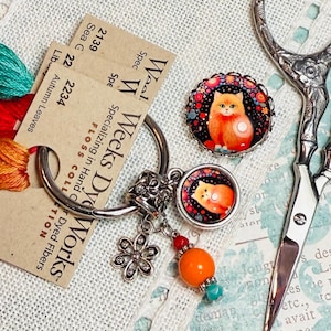 Puede incluir: Un llavero de plata con un dije redondo que presenta una ilustración colorida de un gato. El dije está unido a un anillo de plata con un dije de flor y un borla de cuentas. El llavero se muestra sobre una superficie de tela blanca con carretes de hilo de bordar y un par de tijeras de plata.