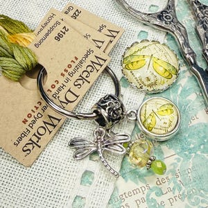 May include: A collection of craft supplies including Weeks Dye Works floss, a dragonfly charm, decorative buttons, and vintage scissors. The floss is green and yellow, and the buttons feature a dragonfly design.
