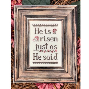May include: A framed cross-stitch embroidery with the text "He is risen just as He said". The embroidery is in a rustic wooden frame with a distressed finish.
