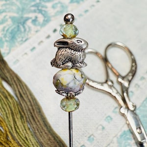 May include: A silver needle minder with a bunny charm, green and yellow beads, and a silver top. The needle minder is next to a pair of silver scissors and green and yellow embroidery floss.