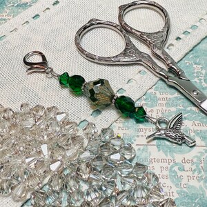 Puede incluir: Un charm de tijera plateado con cuentas verdes y doradas, un charm de colibrí plateado y un cierre plateado. El charm está sobre un fondo de tela blanca.