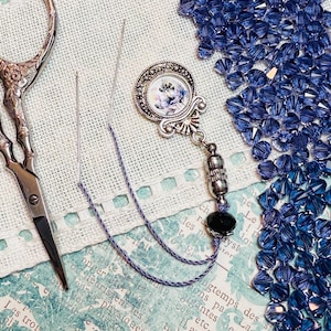 May include: An assortment of craft supplies including ornate scissors, a needle, and a decorative needle minder with a floral design. A pile of blue crystal beads and a length of blue thread are also present. The background features printed paper.