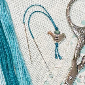 May include: Two silver needles, a skein of blue and white embroidery floss, and a pair of silver embroidery scissors with floral designs. A small blue bird charm hangs from a blue thread.