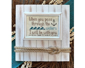 May include: A white wooden frame with a rope border, featuring a cross-stitch design with the text "When you pass through the waters I will be with you - Isaiah 43:2" and an anchor and waves.