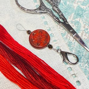 Puede incluir: Un imán para agujas naranja y rojo con un cierre plateado, junto a hilo de bordar rojo y tijeras vintage. El imán para agujas tiene un diseño circular naranja con un patrón metálico. Las tijeras son plateadas con detalles florales.