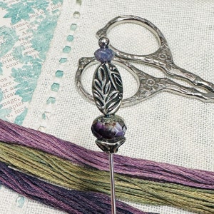 May include: A silver needle with a purple and green gemstone bead and a silver leaf design charm. The needle is lying on a white fabric surface with purple, green, and brown embroidery floss.