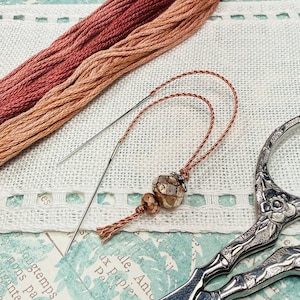 May include: Close-up of embroidery supplies including two skeins of thread in shades of red and peach, a needle threader with a decorative bead, and ornate silver scissors. The items are arranged on a white fabric with a decorative border.