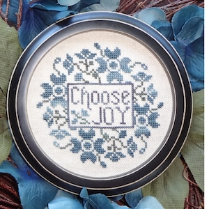 Può includere: Un disegno a punto croce incorniciato con le parole "Choose JOY" in un riquadro rettangolare, circondato da ricami floreali blu. La cornice è nera e lo sfondo presenta un fogliame blu e verde.