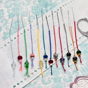 May include: A set of 12 embroidery needles with colorful threads and beads attached. The needles are arranged on a white fabric background with a floral pattern.