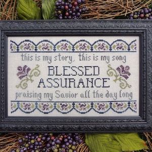 May include: A framed cross-stitch embroidery with the text "This is my story, this is my song Blessed Assurance praising my Savior all the day long". The embroidery features a floral design with purple and green accents.