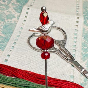 May include: A silver bird charm with red glass beads on a needle, used for needlepoint or embroidery.