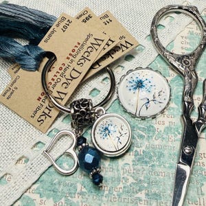 May include: A collection of craft supplies including blue thread, a keychain with a heart charm and a dandelion design, and ornate scissors. The keychain has a blue bead and a round pendant with a dandelion image. The scissors are silver.