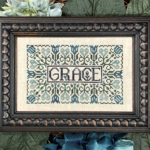 Puede incluir: Un bordado de punto de cruz enmarcado con un diseño floral y la palabra "GRACE" en el centro. El marco es de madera marrón oscuro con un borde de cuentas.