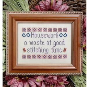Könnte beinhalten: Ein gerahmter Kreuzstich-Sampler mit einem braunen Holzrahmen. Das Kreuzstich-Design zeigt den Text "Housework a waste of good stitching time" mit einem blauen Unendlichkeitszeichen auf beiden Seiten des Wortes "Housework".