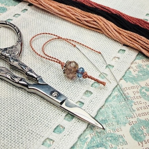 May include: Silver-toned metal embroidery scissors with an ornate floral handle design, a sewing needle threaded with copper-colored embroidery floss, and two decorative beads in brown and blue, alongside strands of black and copper thread.