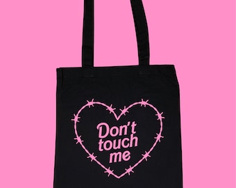 Puede incluir: Bolso tote negro con un diseño de corazón de alambre de púas rosa y el texto "Don't touch me". El bolso tiene dos asas negras y está sobre un fondo rosa.