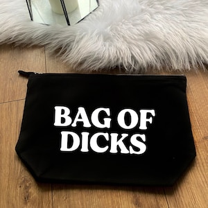 May include: Black zippered pouch with the text "BAG OF DICKS" in bold white letters. The pouch is rectangular and sits on a wooden surface. The background includes a white fluffy rug and a mirror.