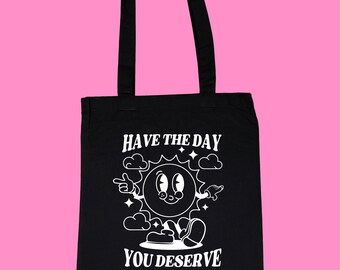 Puede incluir: Bolso tote negro con un gráfico blanco de un sol sonriente, nubes y el texto "HAVE THE DAY YOU DESERVE." El bolso tiene dos asas negras y está hecho de un material resistente. El diseño es de estilo retro de dibujos animados.