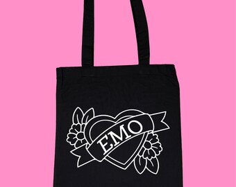 Puede incluir: Bolso tote negro con un gráfico blanco. El gráfico presenta un corazón con una pancarta que dice "EMO", rodeado de flores y hojas. El bolso tiene dos asas negras y está sobre un fondo rosa.