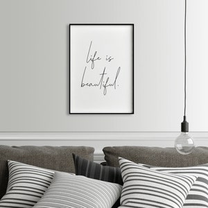 May include: Black and white framed print with the text "Life is beautiful." hanging on a white wall above a gray couch with striped pillows.