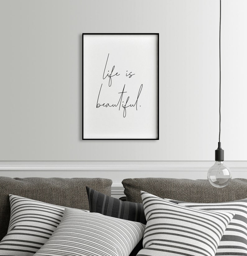 May include: Black and white framed print with the text "Life is beautiful." hanging on a white wall above a gray couch with striped pillows.