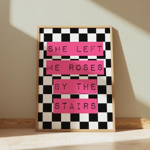Può includere: Stampa artistica incorniciata con sfondo a scacchi bianchi e neri. Il testo "SHE LEFT ME ROSES BY THE STAIRS" è in nero su blocchi rosa. La cornice è di colore legno chiaro.