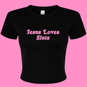 以下が含まれることがあります： 胸の中央に明るいピンク色の丸みを帯びたセリフ体で「Jesus Loves Sluts」というテキストがプリントされた、黒の半袖クルーネックTシャツ。この衣類は、リブ編みのネックラインとストレートな裾を持つスタンダードなフィット感のデザインです。