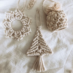 May include: Three handmade macrame ornaments in a cream color. The ornaments are a circle, a tree, and a small pouch with a tassel. The ornaments are hanging on a white fabric background.