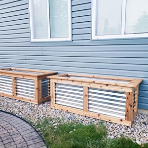 Puede incluir: Dos jardineras de madera con frentes de listones met&aacute;licos, colocadas contra una pared de revestimiento azul. Las jardineras est&aacute;n hechas de madera natural y tienen un aspecto r&uacute;stico.