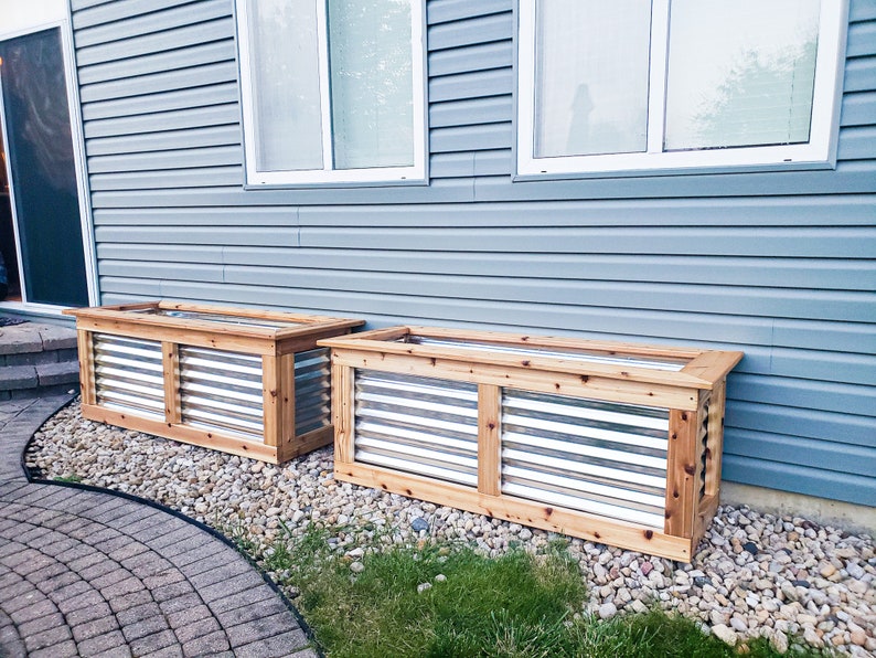 Puede incluir: Dos jardineras de madera con frentes de listones met&aacute;licos, colocadas contra una pared de revestimiento azul. Las jardineras est&aacute;n hechas de madera natural y tienen un aspecto r&uacute;stico.