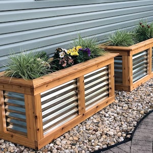 Puede incluir: Dos jardineras de madera con paneles de metal corrugado, llenas de plantas verdes y flores de colores, se colocan sobre una cama de grava frente a una casa con revestimiento gris.