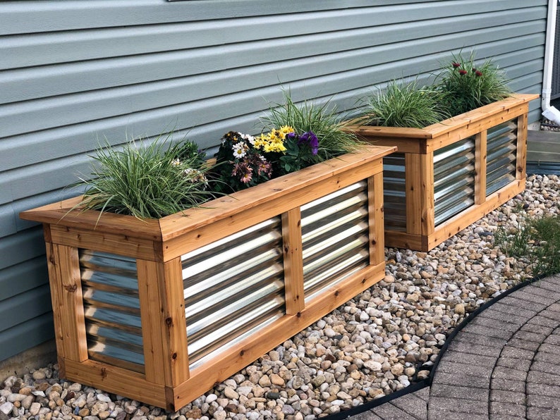 Puede incluir: Dos jardineras de madera con paneles de metal corrugado, llenas de plantas verdes y flores de colores, se colocan sobre una cama de grava frente a una casa con revestimiento gris.