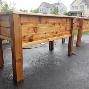 May include: Two wooden raised garden beds with a natural wood finish. The beds are made of cedar planks and have a simple design. The beds are sitting on a black asphalt driveway.