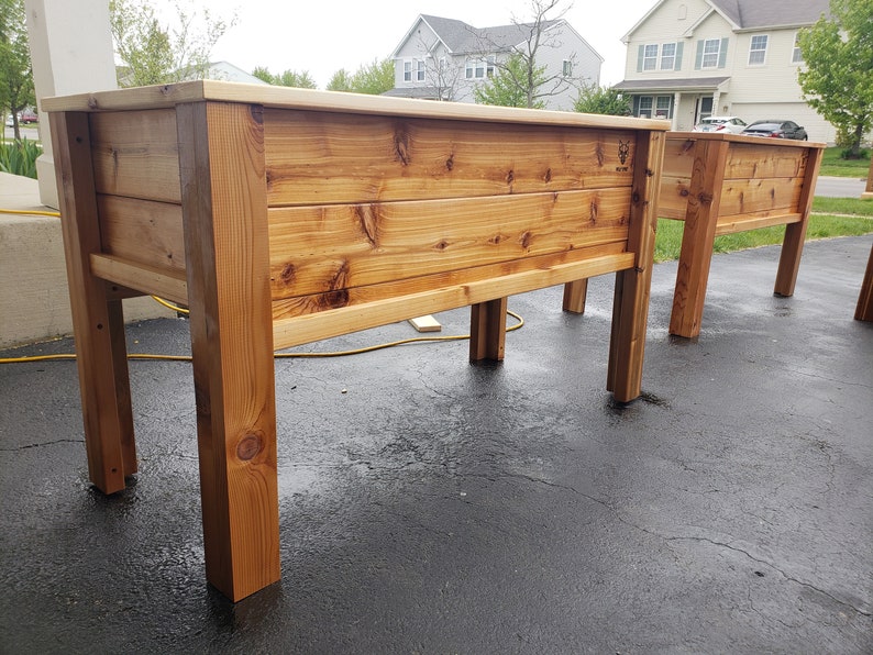 May include: Two wooden raised garden beds with a natural wood finish. The beds are made of cedar planks and have a simple design. The beds are sitting on a black asphalt driveway.