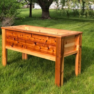May include: A raised wooden garden bed with a natural cedar finish. The bed is elevated on four legs and has a rectangular shape.