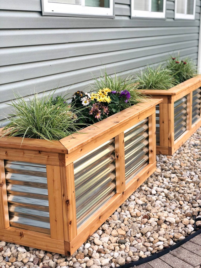 Puede incluir: Dos jardineras de madera con paneles de metal corrugado y una variedad de flores en flor. Las jardineras est&aacute;n sentadas sobre un lecho de peque&ntilde;as piedras blancas.