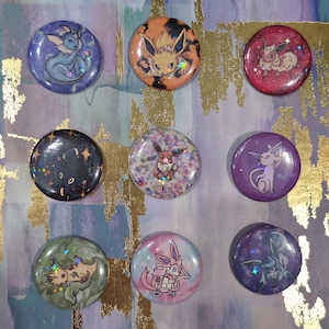 May include: Nine round pinback buttons featuring various colorful illustrations of Pokémon characters. The buttons have a glossy finish and are set against a background with gold accents. The designs include characters like Vaporeon, Flareon, and Eevee.