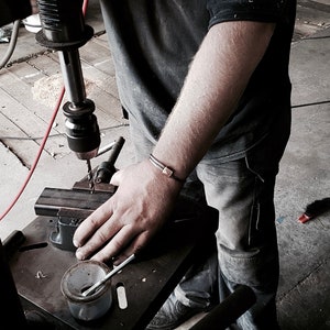 Pode incluir: Uma pessoa usando uma furadeira de coluna para fazer um furo em um objeto de metal. A furadeira de coluna é preta e tem um cabo vermelho. A pessoa está vestindo uma camisa preta e calças jeans azuis. A pessoa está usando uma pulseira no pulso.