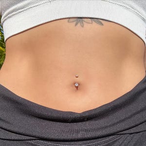 May include: A close-up of a belly button piercing with a small, clear gem. The piercing is centered on the abdomen, with a matching small gem above. The person is wearing a gray sports bra and black athletic pants.