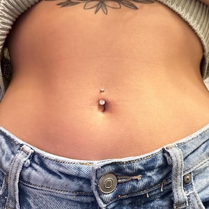 May include: Close-up of a belly button piercing with a sparkling gem. The piercing is centered on the abdomen, with a small stud above and a larger gem below. The person is wearing blue denim jeans.