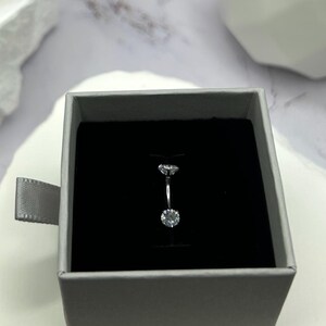 May include: A silver belly ring with two clear cubic zirconia stones. The ring is in a small gray box with a black interior.
