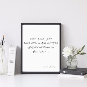 Op de afbeelding: Een ingelijst print met een witte achtergrond en zwarte tekst die "Isn't that just kick-you-in-the-crotch spit-on-your-neck fantastic?" luidt. De tekst is in handschrift geschreven. De print ligt op een wit oppervlak met een vaas met witte bloemen, een boek en een pot met kwasten.