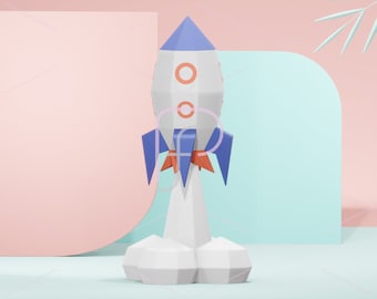 May include: A white, blue, and orange paper rocket ship model on a light blue and pink background. The rocket has two red circles on the side and a blue pointed top. The rocket is sitting on a white base.